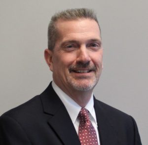 Man with gray scruff smiles at the camera in a black blazer, white shirt, and red tie