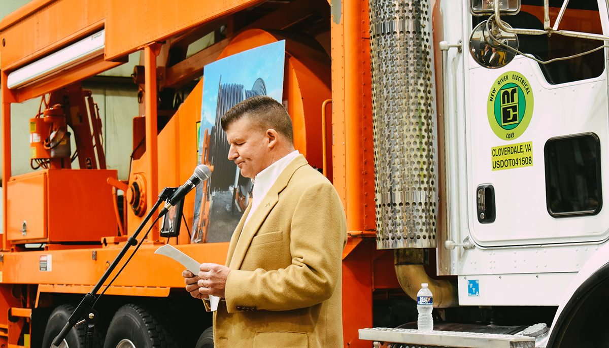 A man in a tan jacket is reading from a paper at a microphone stand, with a large orange construction vehicle and a New River Electrical Corp truck in the background.