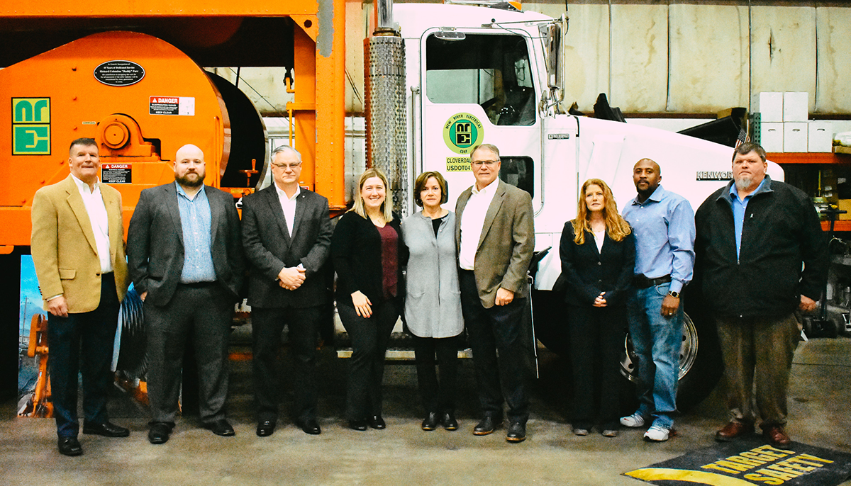 A group of individuals standing in front of an orange construction vehicle and a white truck, both with the New River Electrical Corp logo.