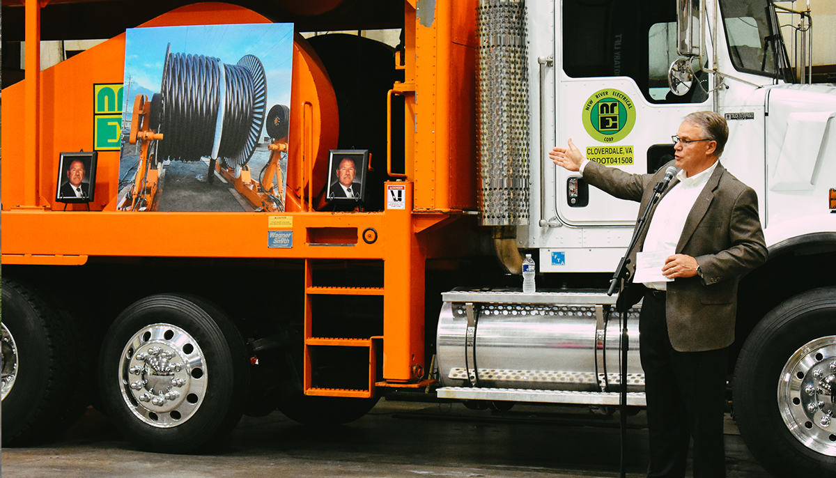 A man in a suit speaking at a podium, gesturing towards an orange construction vehicle with images and a framed photo displayed on it.