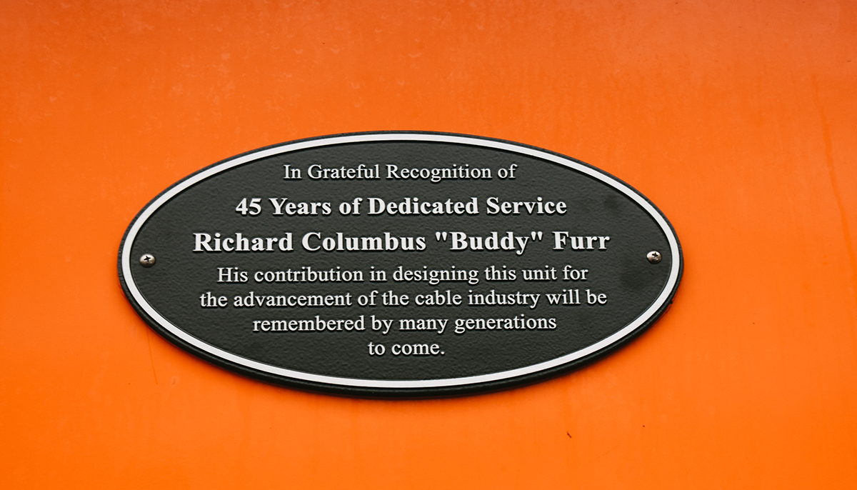 A commemorative plaque on an orange background that reads 'In Greatful Recognition of 45 Years of Dedicated Service, Richard Columbus Buddy Furr, His contribution in designing this unit for the advancement of the cable industry will be remmebered by many generations to come.'