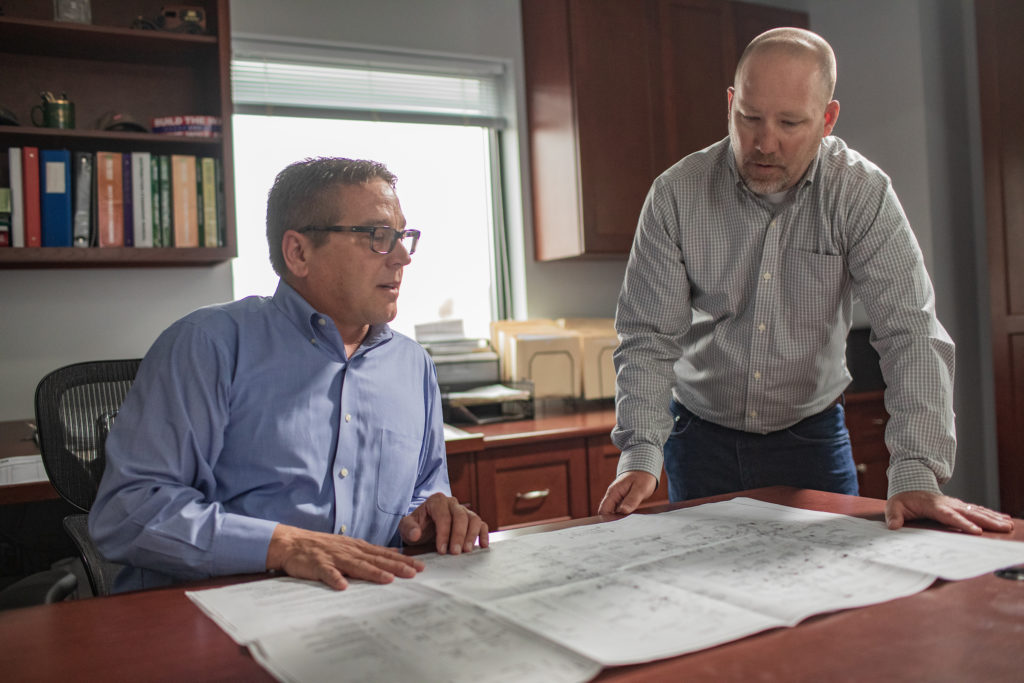 Two men in business casual attire are examining and discussing large architectural plans spread out on a table in an office setting.