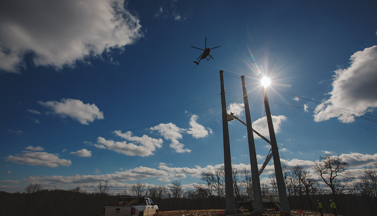 A silhouette of a helicopter flying above utility workers and poles with the sun shining brightly in the background.