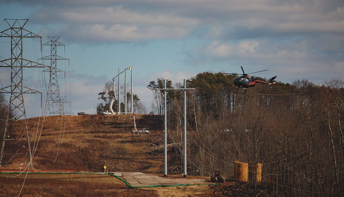 A helicopter is flying near newly installed utility poles in a field with existing high-voltage towers in the background.