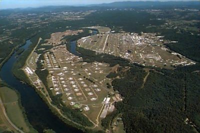 Aerial view of a large industrial facility with multiple buildings and storage units near a winding river, surrounded by forested areas.