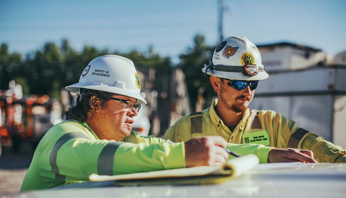 Two New River Electrical employees discussing plans and writing on a notepad on top of truck roof.