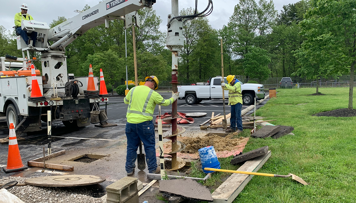 Utility workers in high-visibility clothing are operating a drill from a truck to work on underground installations at a site with traffic cones and exposed earth.