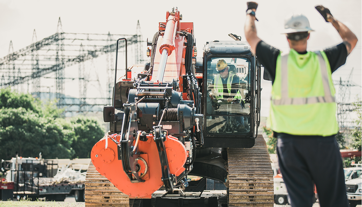 A construction worker in a high-visibility vest is guiding heavy machinery operated by another worker at a construction site.