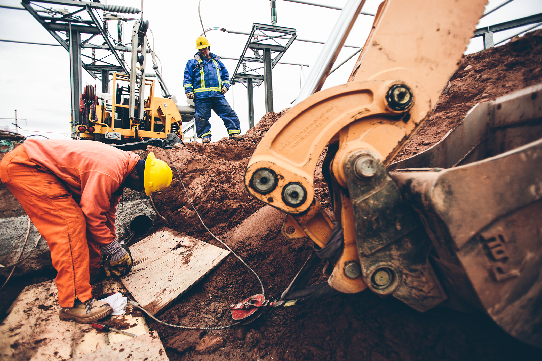 Two New River crew members working with cabling in a dirt hole with an excavator scoop next to them.