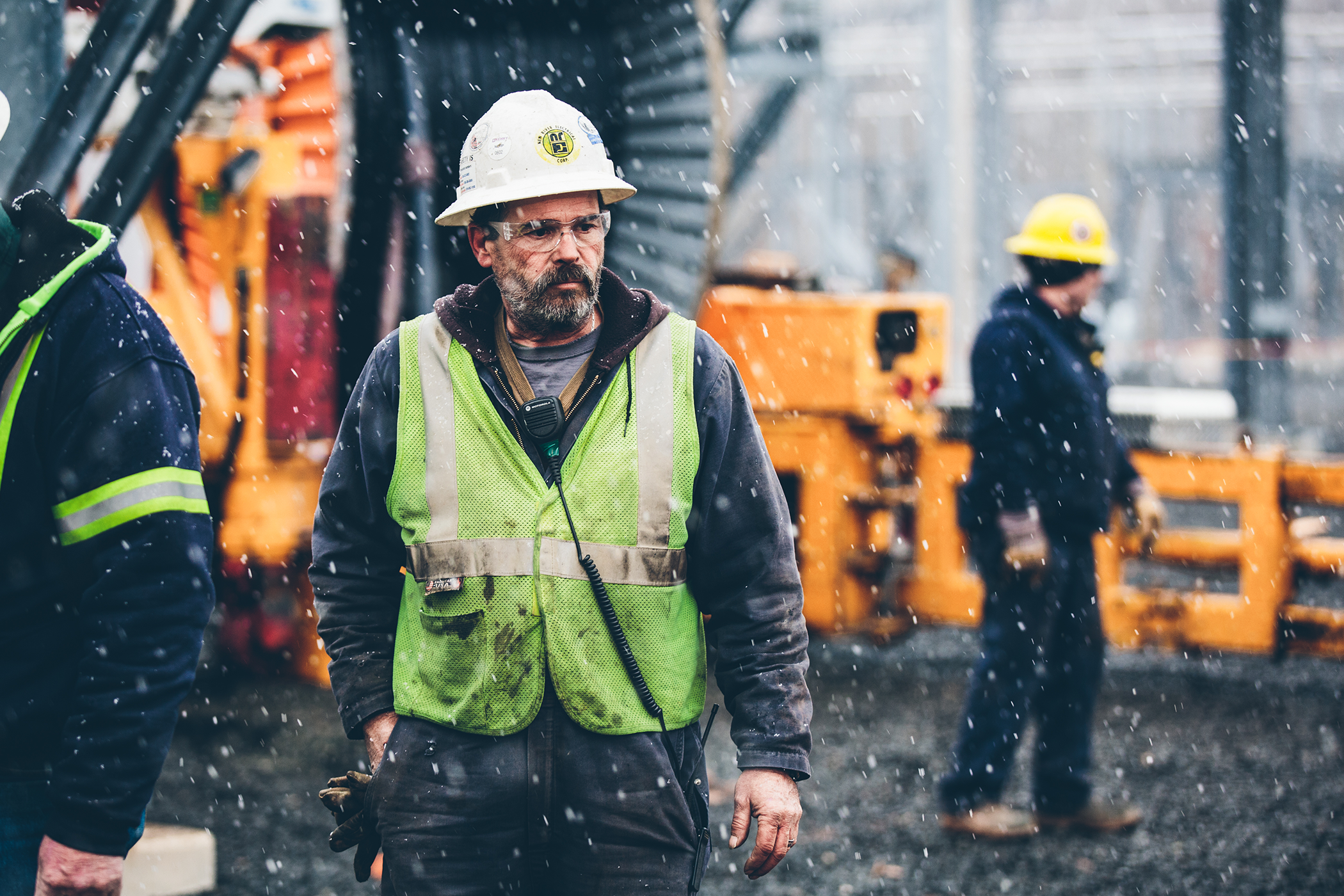 A worker in a reflective vest and hard hat walks through a construction site with heavy machinery in the background, under a light snowfall.