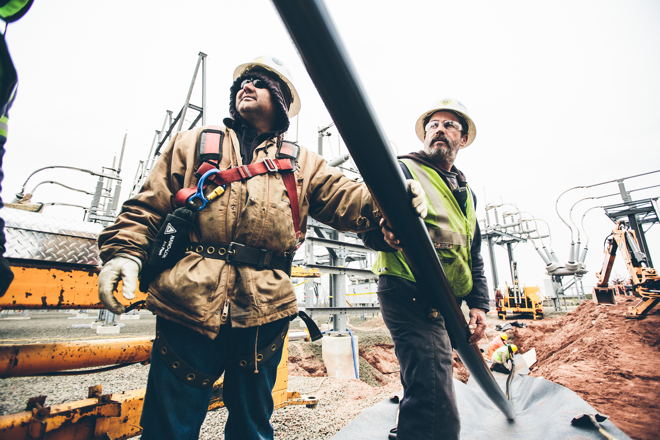 Two workers in protective gear and hard hats are handling piping equipment at an industrial site with complex machinery in the background.