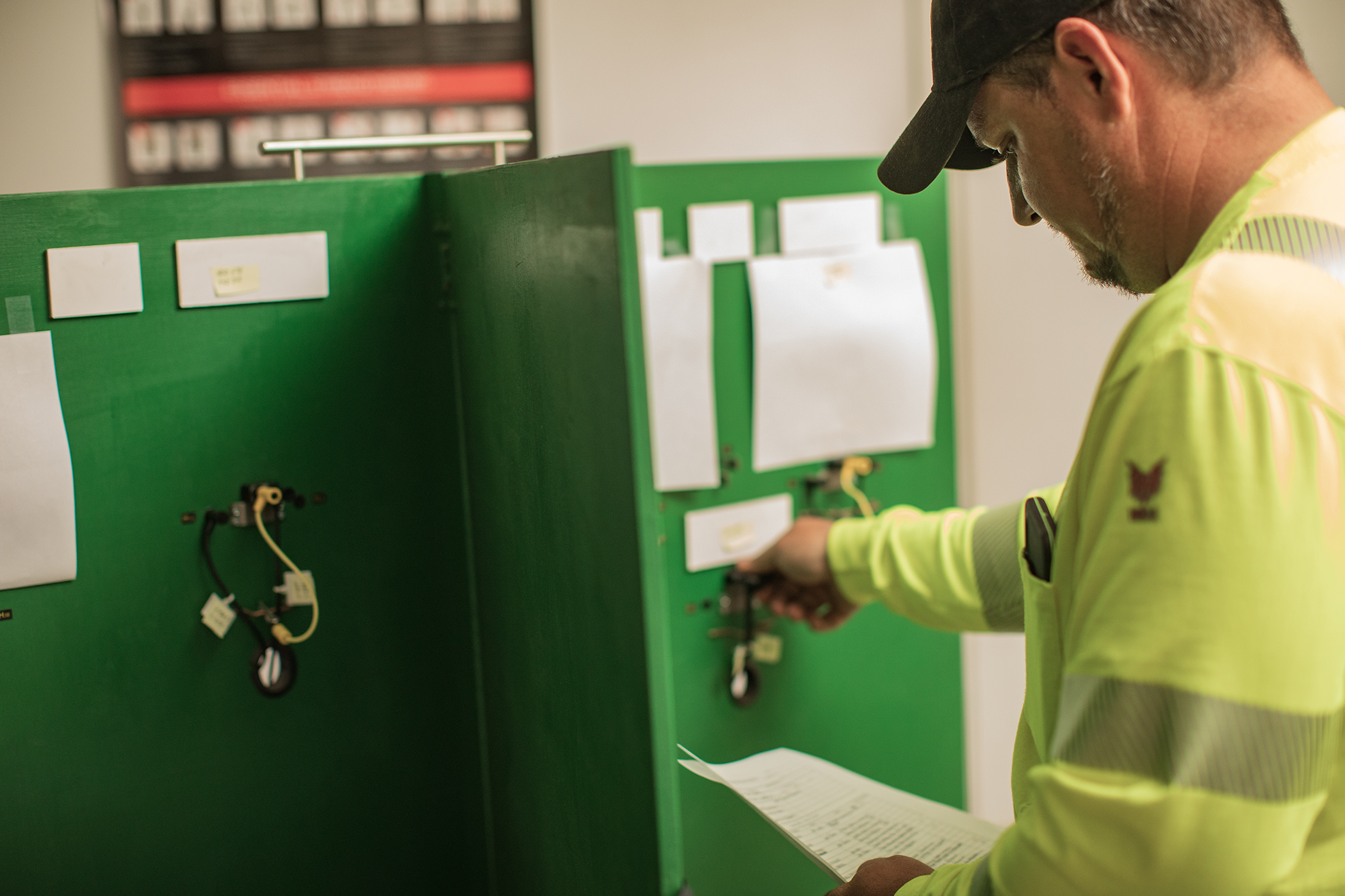 Man in yellow safety shirt looking at a piece of paper and adjusting wires attached to a wooden structure painted green.