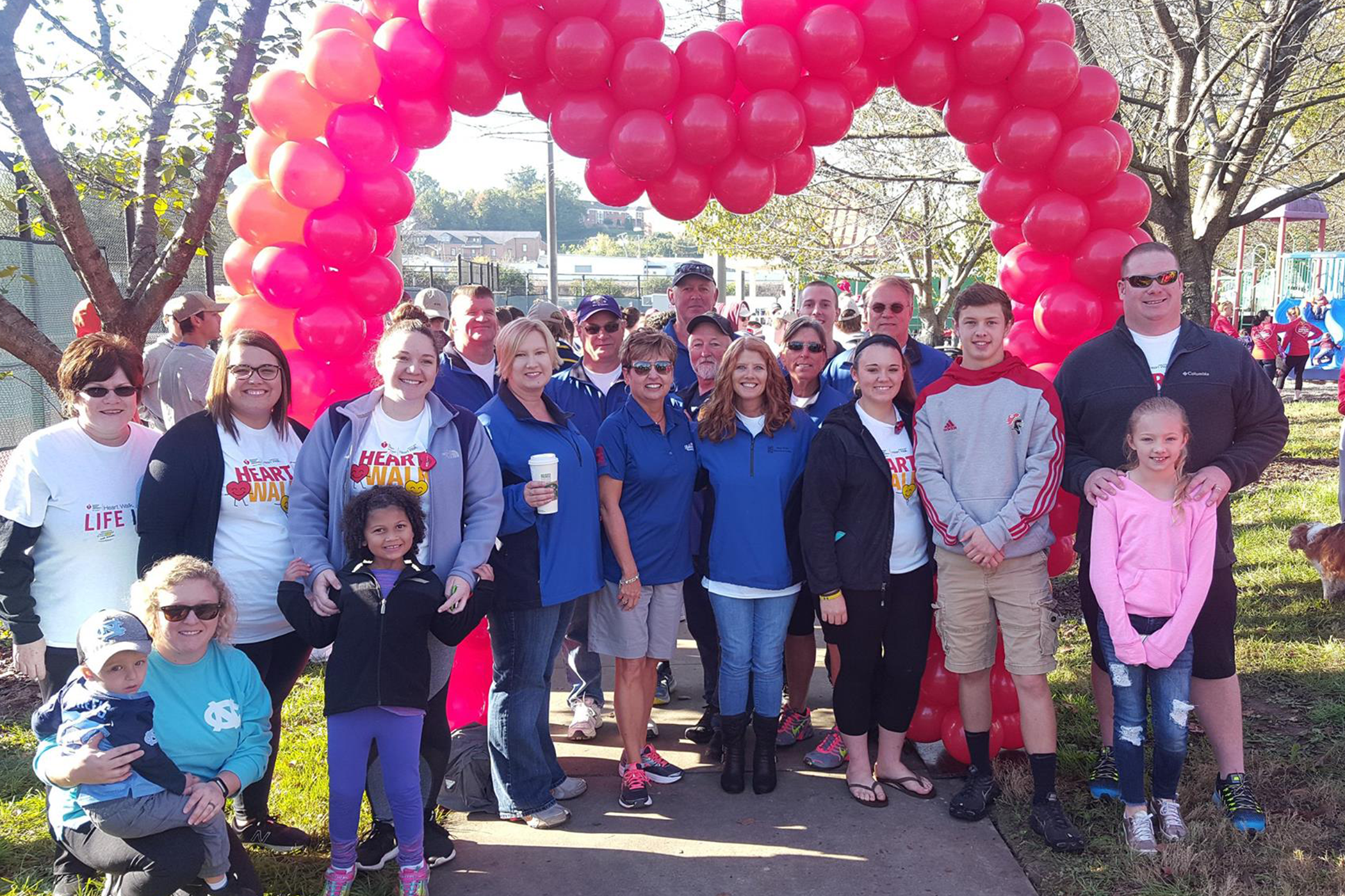 Group shot of men, women, and children posing beneath a heart-shaped red balloon arch in a park.