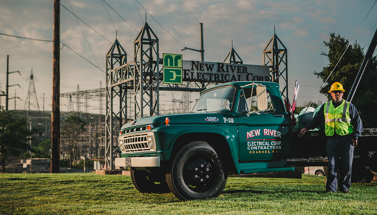 An original green New River Electrical truck with an employee in equipment standing in front of a New River Electrical sign and substation.