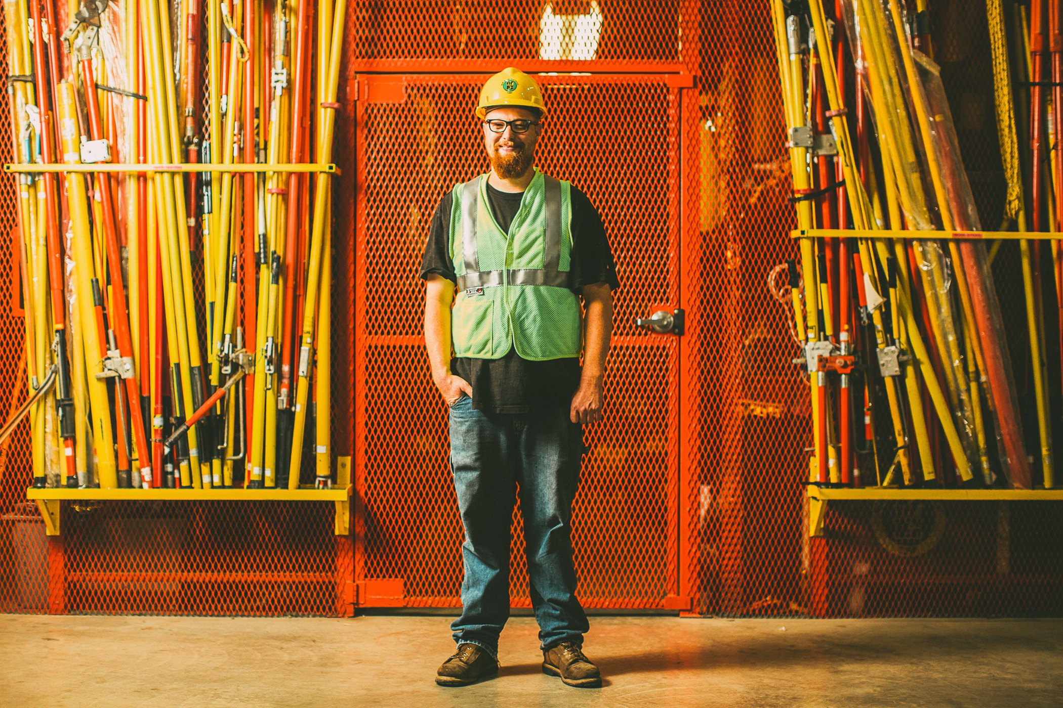 NRE employee in safety equipment standing in front of a grated orange wall with yellow construction poles standing behind them.