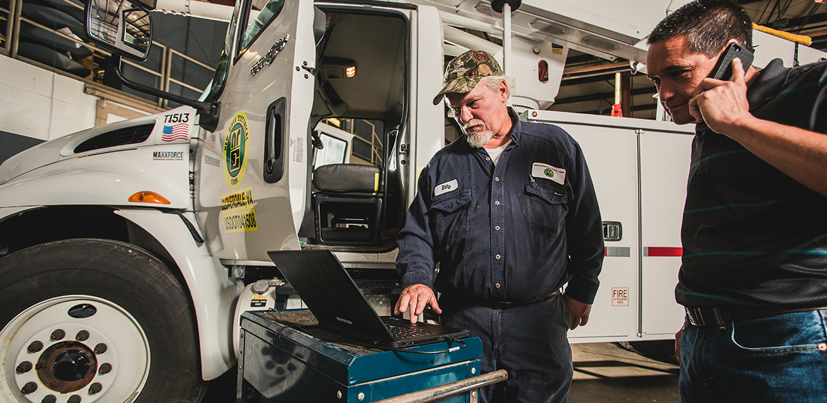 Two New River Electrical operations department team members studying data on a computer while the other is on the phone in front of a New River bucket truck.