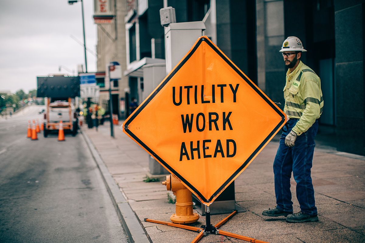 A NRE employee standing next to a Utility Work Ahead safety sign on a roadway job site.