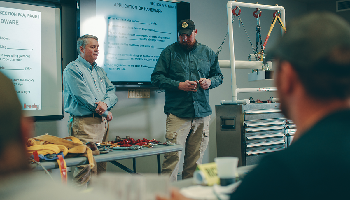 Two men are presenting in a training session with lifting equipment on the table in front of them and rigging hardware instructions displayed on a screen behind.