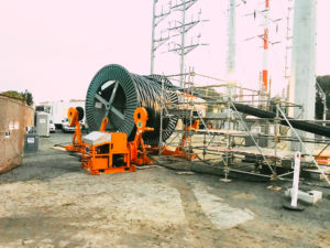 An industrial cable reel and machinery on a construction site with scaffolding and electrical towers in the background.
