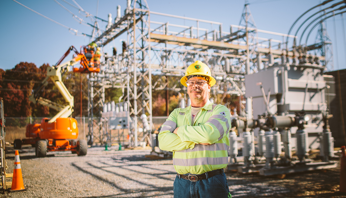 A New River Employee-owner standing with arms crossed in front of a substation and lift.