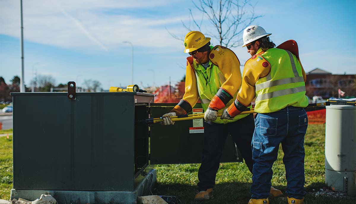 Two New River Electrical employees in yellow and green safety equipment holding a yellow pole into a green electrical box outside.