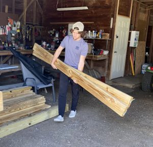 Man in baseball hat carrying two large wooden boards in a workshop.