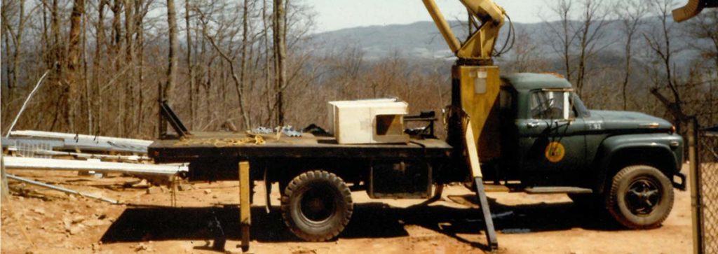 A green and yellow NRE crane truck from 1981 at an outdoor job site.