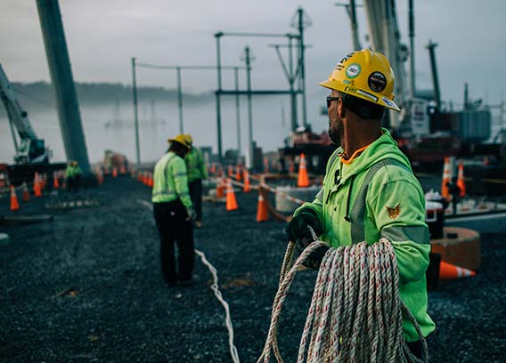 A New River Employee in safety equipment unwinding rope to other employees at a substation job site.