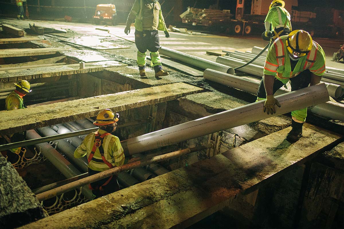 Workers in reflective vests and hard hats are working at night on a construction site with exposed underground piping.