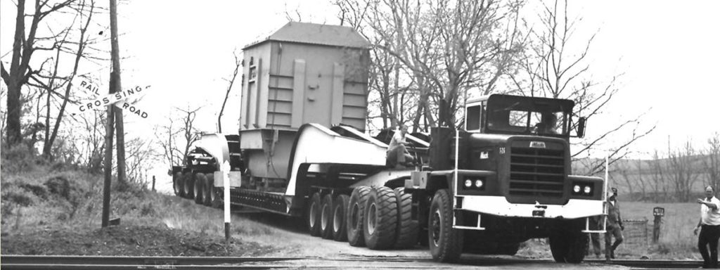 A black and white photo of a large truck and trailer moving a 765kv transformer for a rigging job across the railroad.