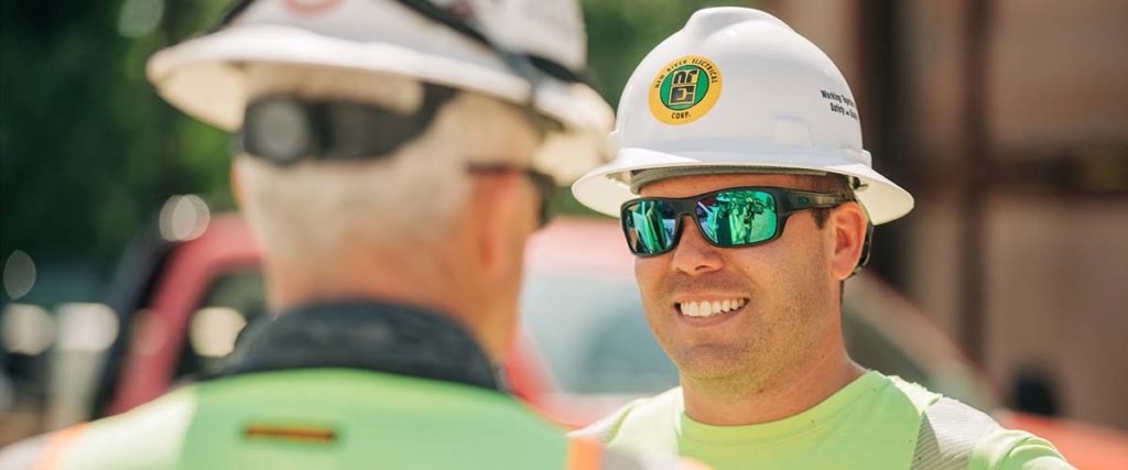 Two New River Employees looking at each other with sunglasses and white hardhats with the NRE logo on them.