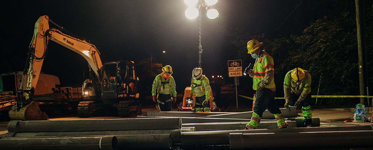 NRE workers arranging cylindrical pipes at night beneath floodlights.