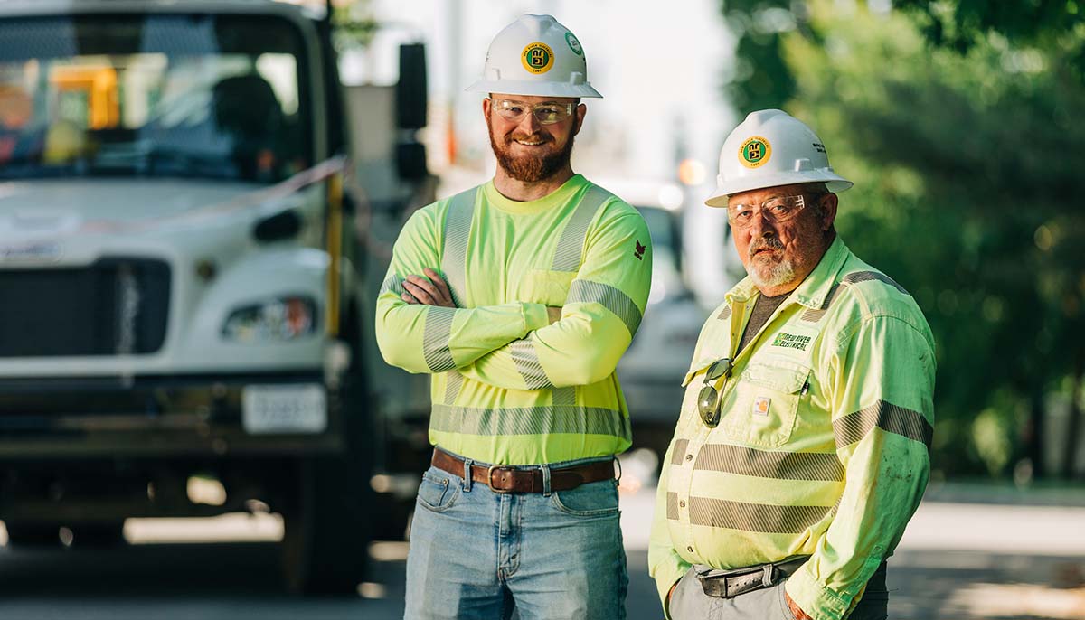Two New River Electrical employees in work vests and hard hats smiling in front of a truck.