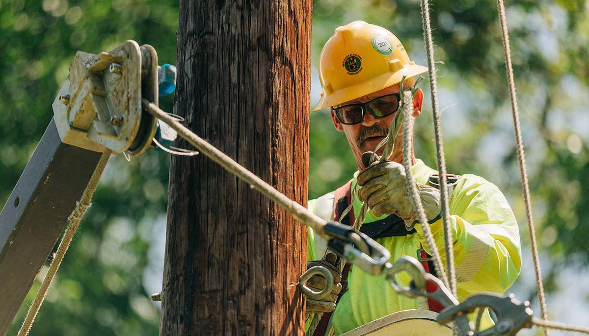A New River Lineman with a yellow hard hat working on an electrical pole while rigging cables.