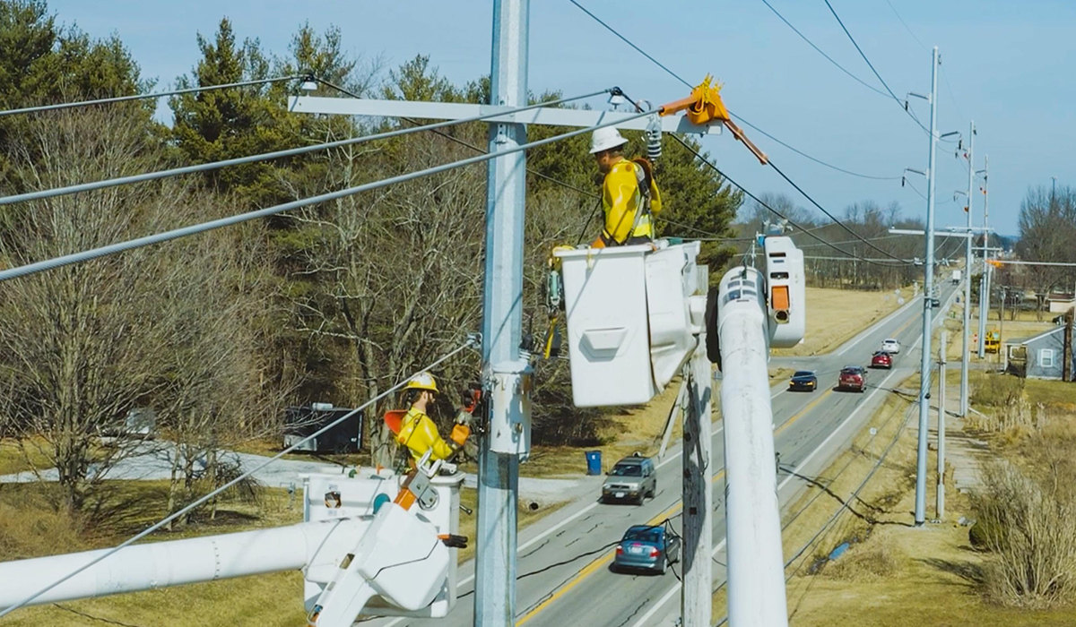 Two utility workers in elevated bucket trucks are working on power lines beside a road with traffic.