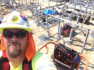 A selfie of Mark Nottingham wearing a hard hat with stickers and a high-visibility vest, with an industrial construction site and equipment in the background.