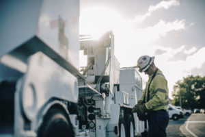 A construction worker in a reflective vest and hard hat is inspecting equipment on a utility vehicle, with the sun low in the sky behind him.