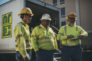 Three construction workers in high-visibility clothing and hard hats are engaged in a discussion beside a truck with a company logo.