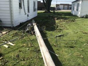 A fallen utility pole with attached wires is lying across a residential lawn, with debris scattered around, indicating recent storm damage or an accident.