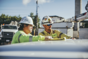 Two workers wearing reflective vests and hard hats review plans on the hood of a truck at a construction site.