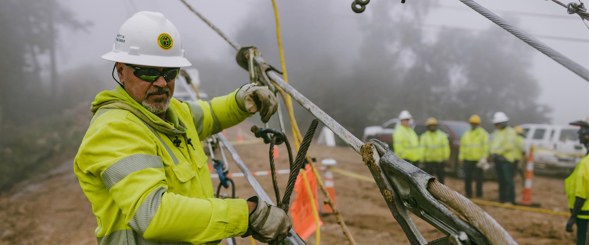 Man in yellow safety jacket, glasses, gloves, and hard hat works in the foreground while group of similarly-attired workers are blurred in the background