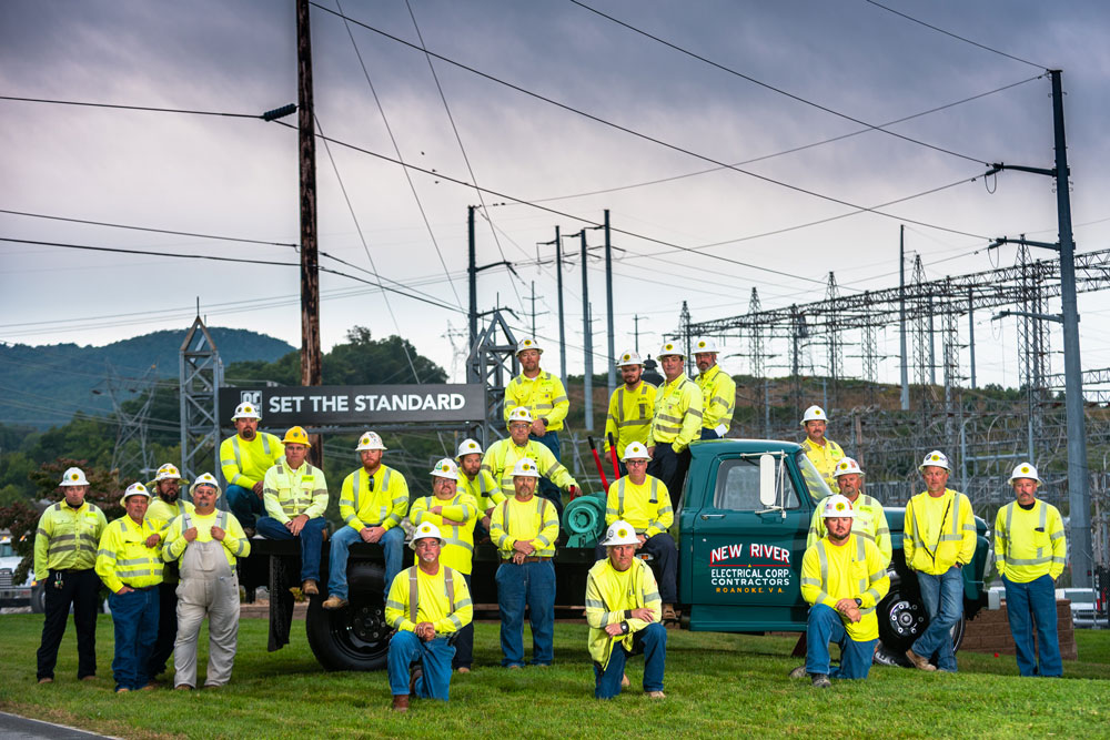 Group of NRE workers in yellow safety shirts and hard hats pose for the camera outdoors in front of a green branded pick up truck and a sign that says 