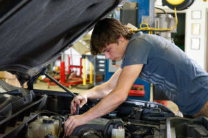 Brown-haired teenaged boy in a gray-blue tshirt leans over the engine of a car with the hood popped.