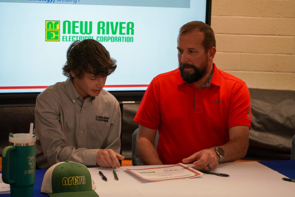 Man in bright red under armor polo shirt sits at a table in front of the NRE logo and awards a certificate to a young brown-haired man in an NRE gray button down