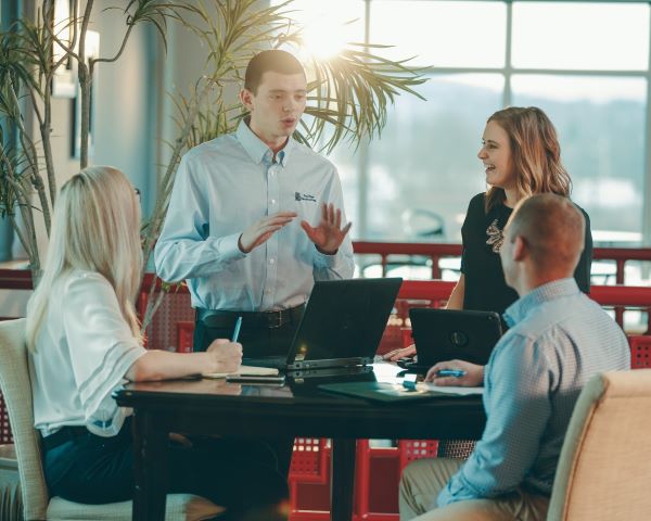 Brown-haired young man in blue button down speaks to another man and two women who are working at a table in front of a large window