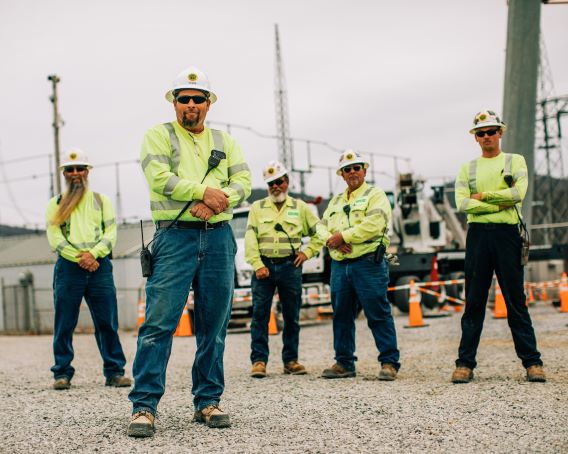 NRE workers in yellow work shirts and hard hats pose for the camera in a gravel lot