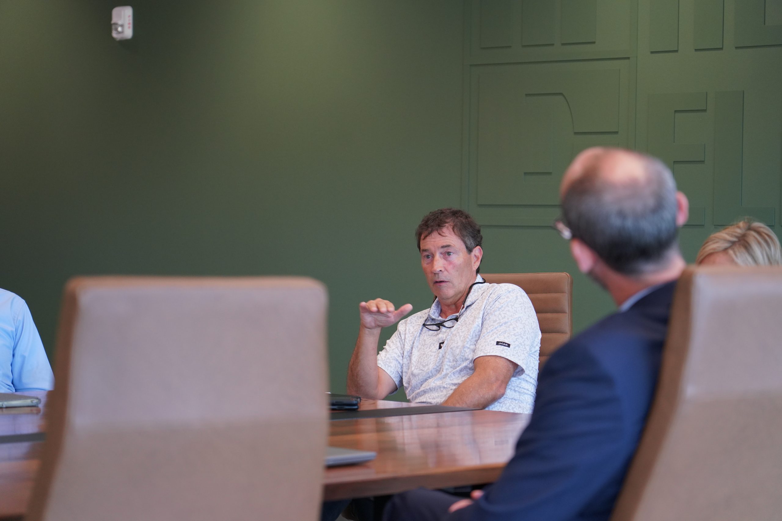 Man in a white short-sleeved collared shirt and black glasses on a landyard around his neck sits at the head of a table and talks in front of a sage green wall