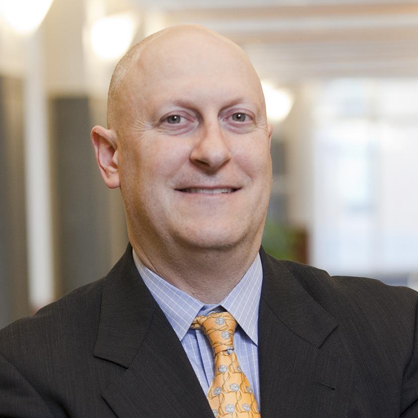 Dan Oglevee, a bald middle-aged man in blue collared shirt, yellow tie patterned with pale blue roses, and black blazer, smiles for the camera