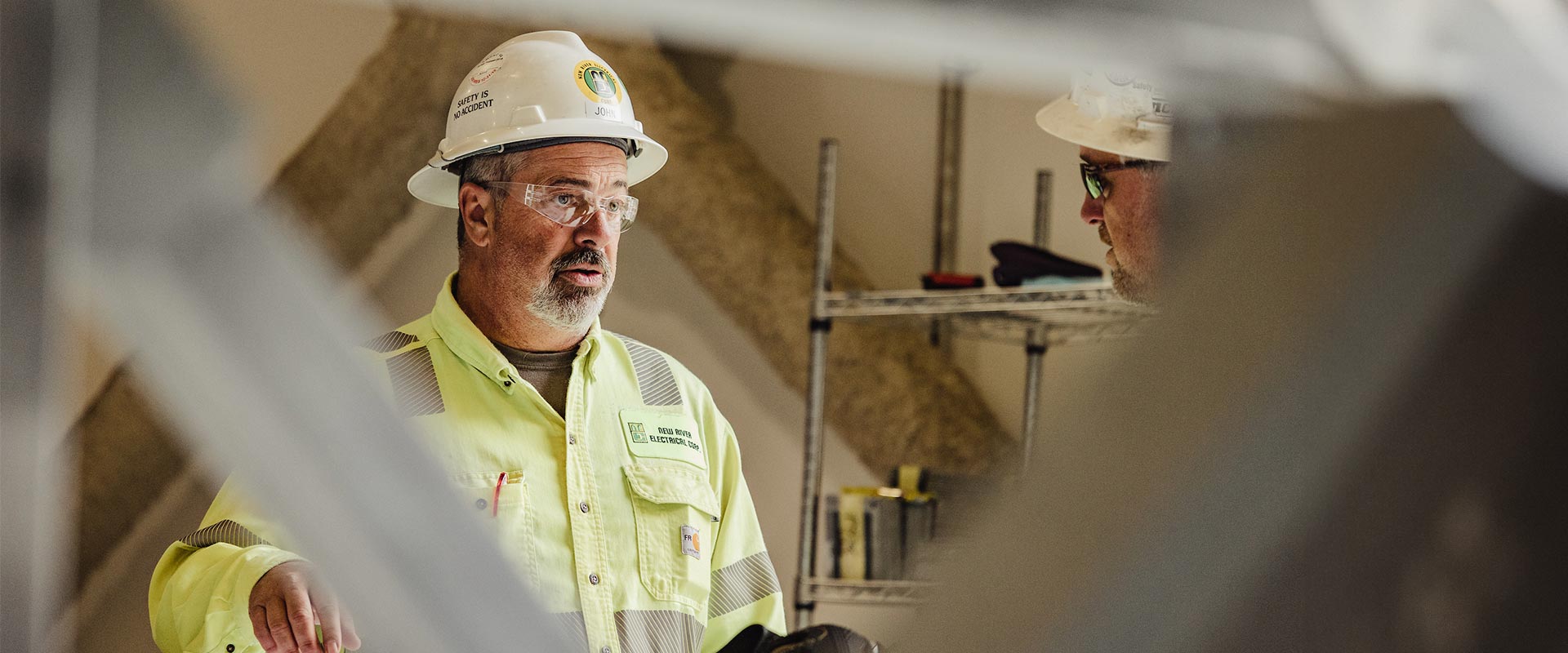 Shot of a man in white hard hat, safety glasses, and yellow safety jacket talks to another worker in sunglasses and white hard hat with blurred white structuring in the foreground.