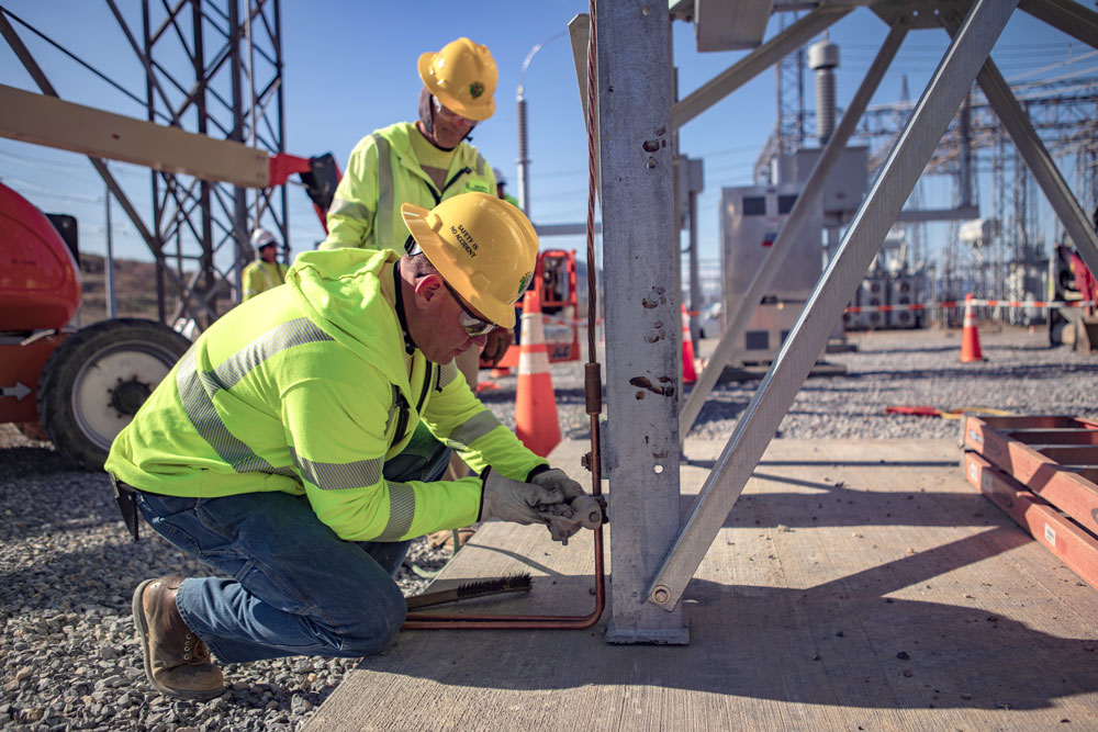 A man in a yellow safety shirt, safety goggles, and yellow hard hat kneels on the gravel and works on a large metal structure while another worker watches from the side